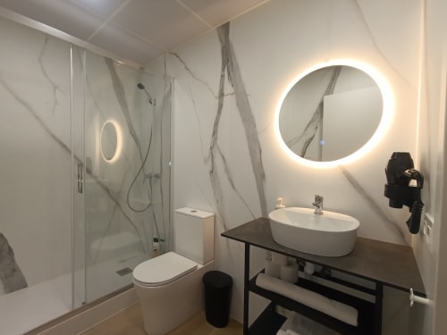 Baño moderno en apartamento El Rincón de Sevilla con paredes de mármol, espejo redondo iluminado, lavamanos blanco y cristal