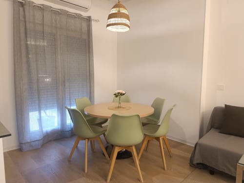 Comedor moderno con mesa redonda de madera, sillas verde salvia, lámpara colgante tejida y cortinas grises en El Rincón de Sevilla