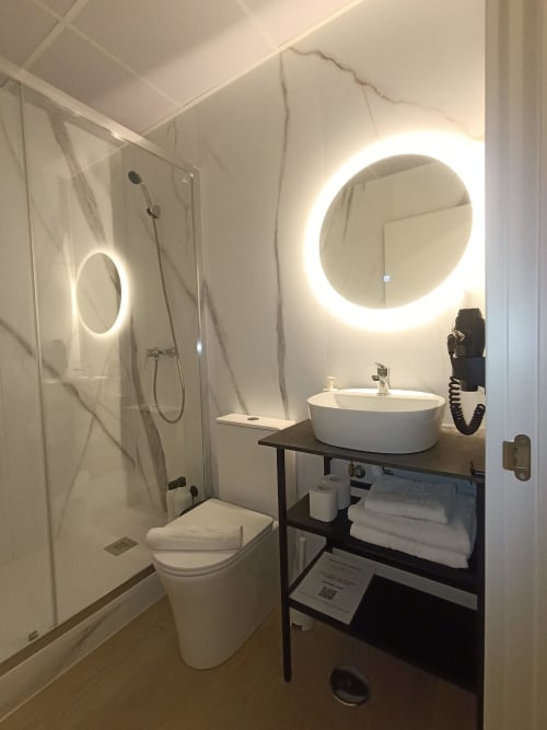 Baño moderno en El Rincón de Sevilla con espejo circular iluminado, lavamanos blanco, inodoro y ducha de cristal