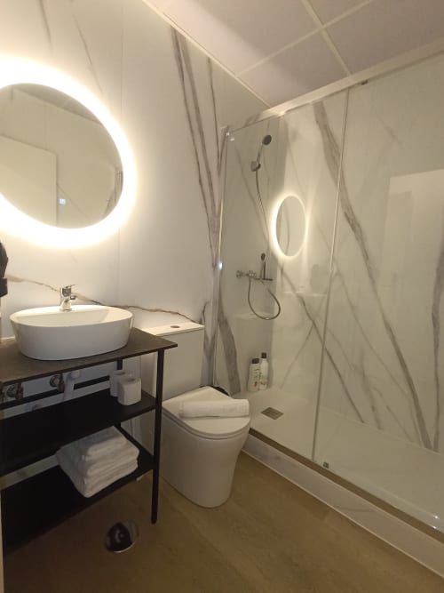 Baño moderno con espejo circular iluminado, lavamanos de cerámica blanca, inodoro y ducha abierta en El Rincón de Sevilla