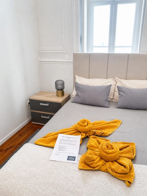 Quarto brilhante de hostel em Lisboa com cabeceira estofada bege, almofadas cinzas, criado de madeira e toalhas amarelo dourado