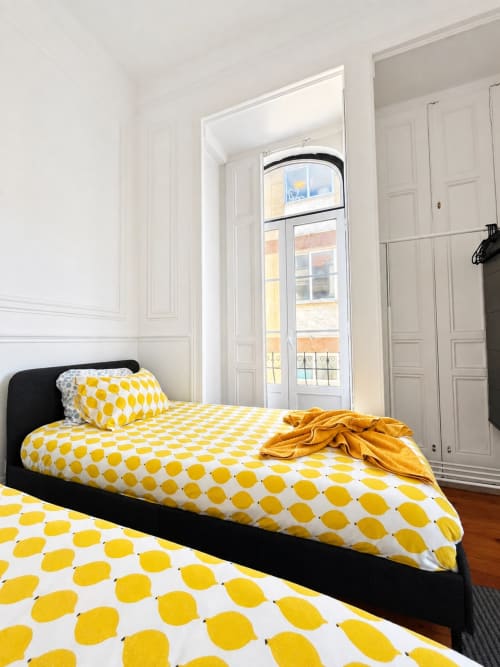 Quarto luminoso em hostel em Lisboa com roupa de cama amarela com poás, cama de estrutura preta e janela alta com vista para a cidade