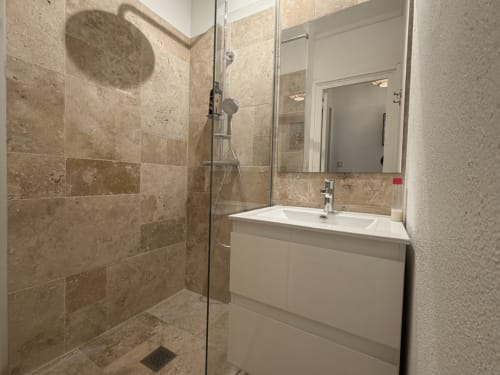 Modern bathroom at Horizon Doré hotel in Gourbeyre with travertine tile, walk-in shower, white vanity, and mirror.