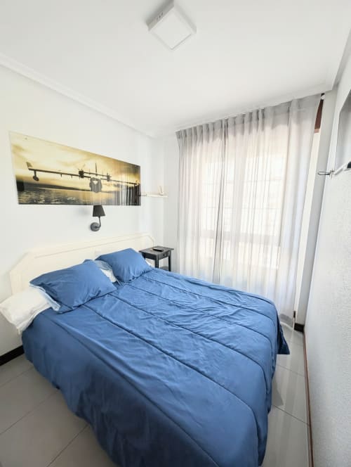 Dormitorio moderno con ropa de cama azul en Alojamiento La Ballena de Santoña, cabecero blanco y cortinas translúcidas