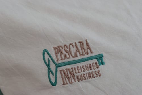 Pescara INN logo embroidered on white fabric with teal and brown stitching details
