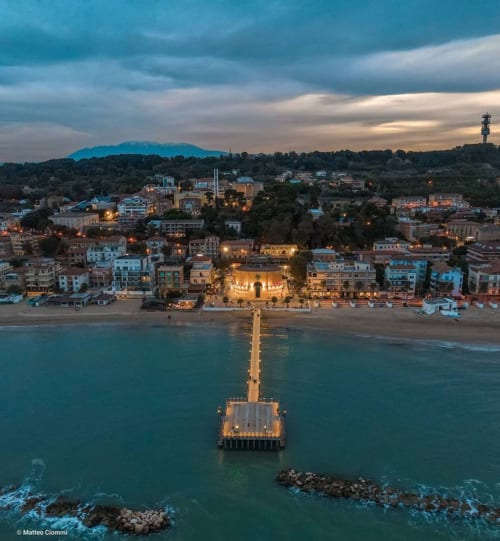Pescara beachfront at dusk with illuminated pier, colorful buildings, and mountains under dramatic sky