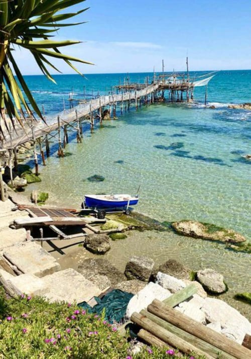 Pescara beachfront with wooden pier, blue fishing boat, clear turquoise water, and pink wildflowers overlooking the Adriatic