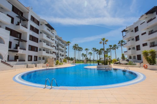 Piscina curva con palmeras y edificios blancos de apartamentos en Haus Holiday Homes, San Pedro del Pinatar