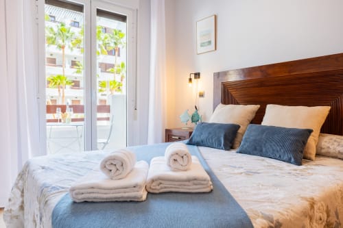 Dormitorio luminoso con camas gemelas, toallas enrolladas, cabecero de madera y vista al balcón de palmeras en Haus Holiday Homes