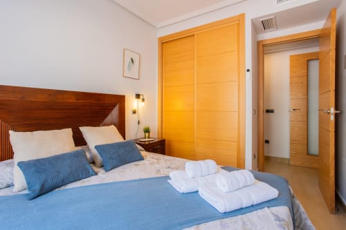 Dormitorio moderno y luminoso con cama de madera, almohadas azules, toallas blancas y armario amarillo dorado en San Pedro del Pinatar