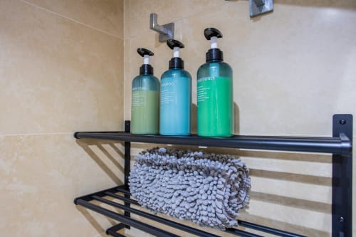 Estante negro de baño con tres botellas dispensadoras (jabón, champú, acondicionador) y paño gris en apartamento Haus Holiday Homes