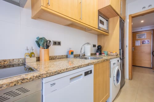 Moderna cocina con encimeras de granito, muebles de madera clara, electrodomésticos blancos y fregadero de acero inoxidable en Haus Holiday