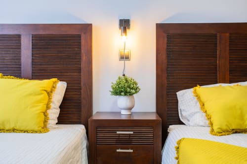 Dos camas individuales con almohadas amarillas y sábanas blancas, cabeceros de madera oscura, mesita central con planta, apliques de pared