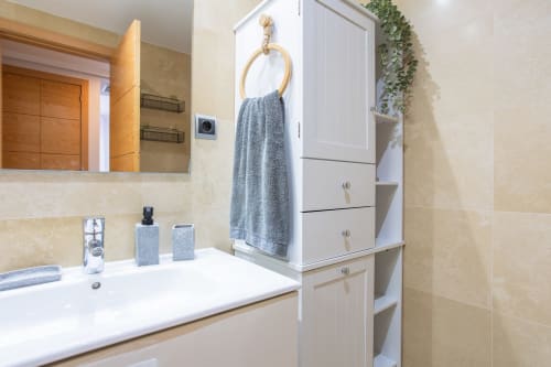 Baño moderno con lavamanos blanco, espejo, toalla gris en perchero de latón, armario blanco con estantes y paredes de azulejos beige