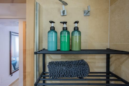 Estante del baño con dispensadores de champú turquesa, acondicionador verde y jabón corporal sobre una toalla de microfibra azul en Haus
