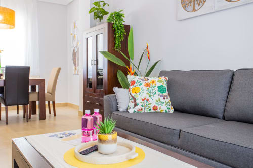 Sala moderna con sofá gris, cojín floral colorido, plantas en maceta y muebles de comedor de madera en Haus Holiday Homes