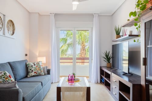 Sala de estar moderna con sofá azul, televisor plano, muebles de madera, plantas en macetas y balcón con vista a palmeras