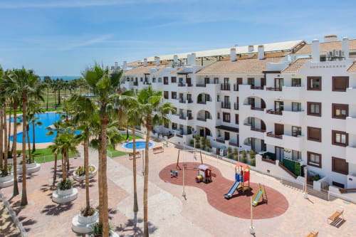 Complejo residencial blanco con piscina, parque infantil y palmeras en Haus Holiday Homes, San Pedro del Pinatar, España