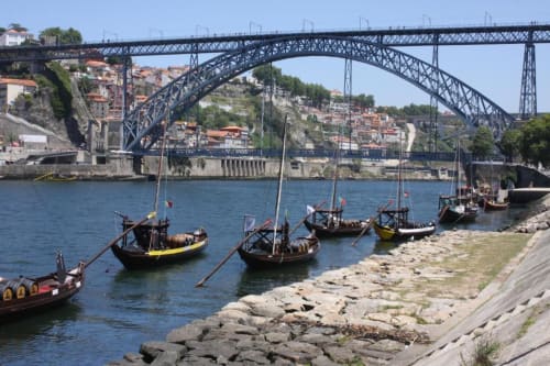 Barcos de madeira tradicionais atracados no Rio Douro com a Ponte Dom Luís e a arquitetura da encosta do Porto ao fundo