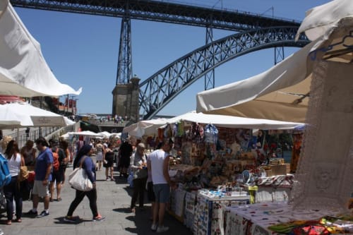Mercado à beira-rio no Porto com ponte icónica, tendas brancas e visitantes explorando os stands dos vendedores