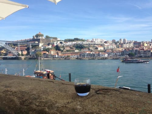 Taça de vinho na Praia da Ribeira, Porto, com o Rio Douro, barcos e arquitetura histórica incluindo a Ponte Dom Luís ao fundo