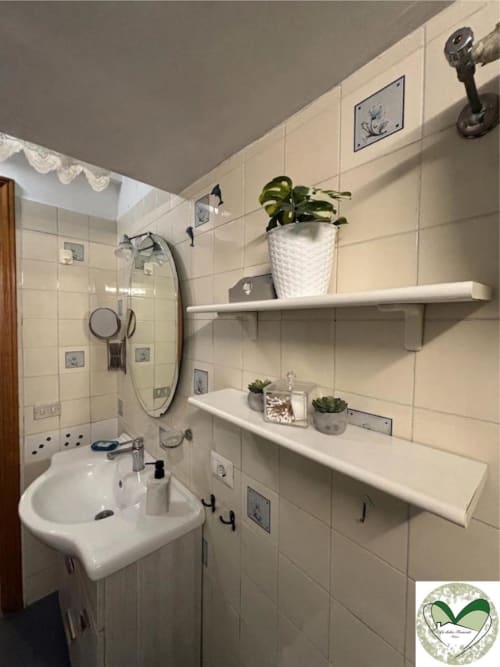 Bagno moderno presso I Dolci Appartamenti Ascani Roma con piastrelle bianche, specchio rotondo, ripiani con piante e oggetti decorativi