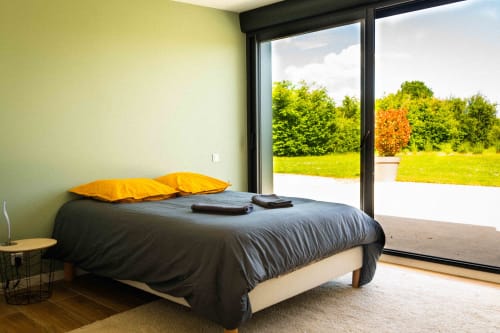 Chambre moderne avec coussins jaunes, literie foncée et grandes fenêtres donnant sur un paysage verdoyant au gîte Accolade