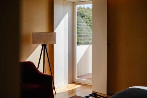 Quarto de hotel moderno no Douro Land com janelas do piso ao teto, luminária de tripé, poltrona bordô e vista para as montanhas da varanda
