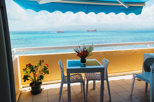 Balcony with sea view at La Perle Fleur D'epée hotel in Le Gosier, featuring blue awning, dining table, and turquoise ocean