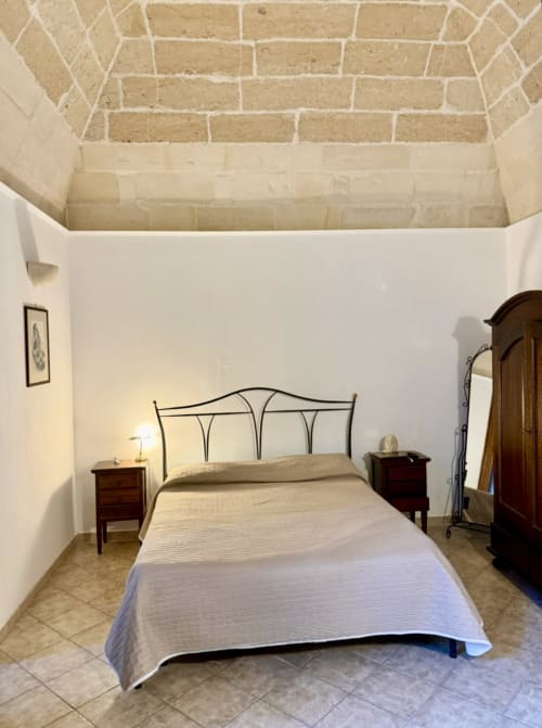Camera luminosa dell'appartamento HABITARE Lecce con letto in ferro, muri bianchi, soffitto in pietra a vista e mobili d'epoca
