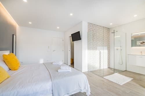 Quarto moderno em apartamento em Albufeira com casa de banho integrada, roupa de cama branca, detalhes em amarelo e base de duche de vidro
