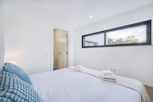 Quarto moderno no BlueHorizon em Albufeira com cama queen, roupas de cama brancas, manta azul, banheiro privado e janela panorâmica