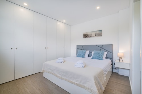 Quarto moderno do apartamento BlueHorizon em Albufeira com guarda-roupas brancos, cabeceira cinzenta e decoração de praia