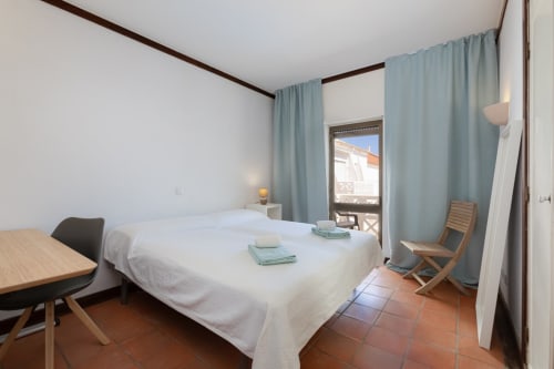 Quarto luminoso no Beachtime Oura em Albufeira com cama branca, piso em terracota e cortinas azuis emolduradas pela vista da varanda