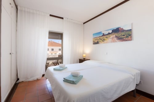 Quarto luminoso de apartamento em Albufeira com cama branca, arte de paisagem desértica, piso em terracota e varanda com vista