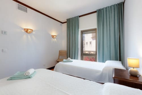 Quarto de hotel luminoso na SignatureStays em Albufeira com duas camas, cortinas menta e vista de varanda