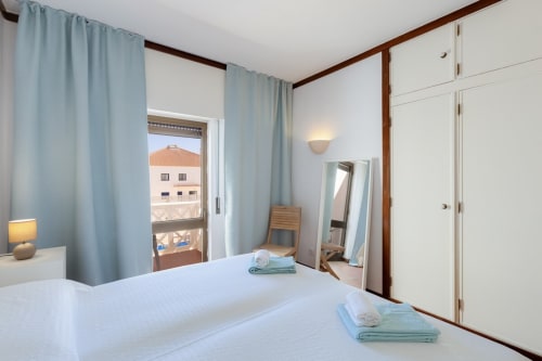Quarto moderno na Beachtime Oura by SignatureStays em Albufeira com varanda vista mar, cortinas azuis e móveis brancos.