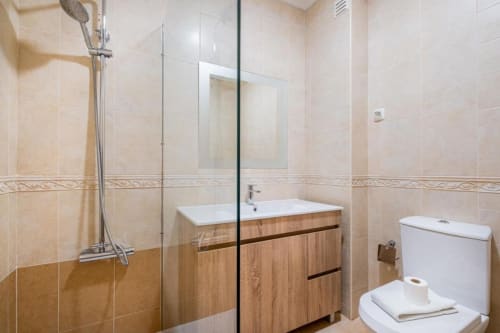 Banheiro moderno em apartamento Farmvillage, Albufeira. Móvel de madeira, box de vidro, vaso branco, azulejos bege.