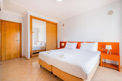 Quarto moderno na Farmvillage by SignatureStays em Albufeira com detalhes em laranja, roupa de cama branca e guarda-roupa espelhado