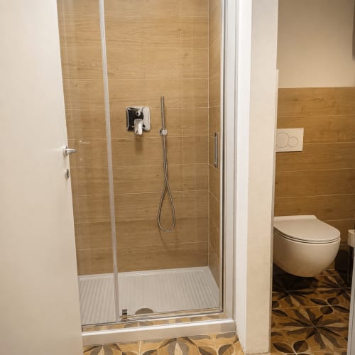Modernes Badezimmer mit begehbarer Dusche, Glasverkleidung, Holzfliesenwände, weißes WC und gemustertem Boden in der Bergamo-Wohnung