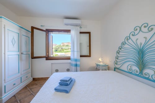 Camera da letto luminosa nella villa di Olbia con accenti turchesi, letto bianco, infissi in legno e vista sulla campagna