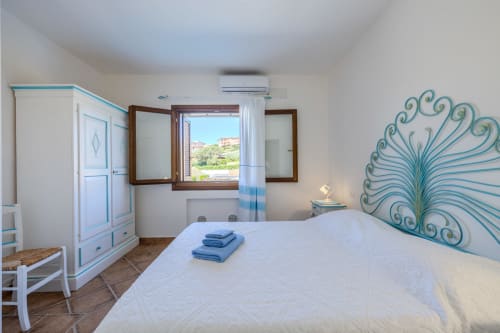 Camera da letto luminosa di villa a Olbia con letto bianco, testata turchese, infissi in legno, climatizzatore e pavimento in cotto