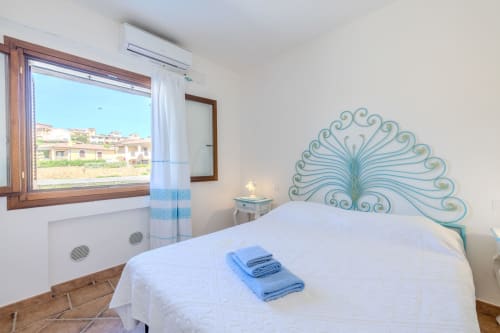 Camera luminosa in villa a Olbia con testata pavone turchese, letto bianco, asciugamani blu, finestra in legno con vista sulla campagna