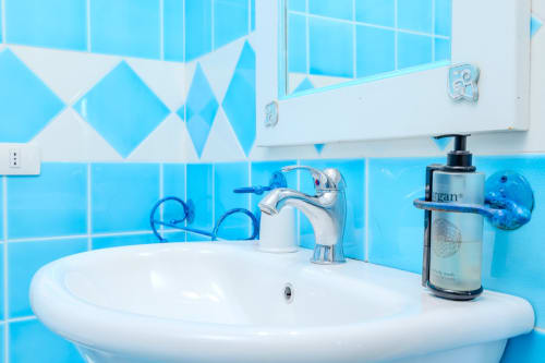 Bagno azzurro luminoso con lavandino bianco, rubinetto cromato e dispenser di sapone nella villa Perseo Case Vacanze a Olba