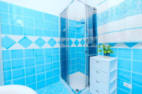 Bagno moderno con piastrelle blu brillanti, box doccia in vetro, vanity bianca e pianta in vaso presso Perseo Case Vacanze
