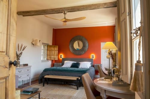 Suite élégante avec mur d'accent orange, lit foncé, poutres en bois et mobilier classique aux Jardins de Baracane