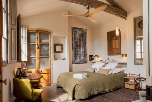 Chambre lumineuse aux Jardins de Baracane à Avignon avec lit, ventilateur en bois, chaise jaune et vue sur le jardin