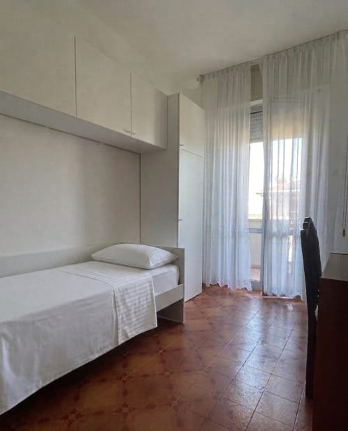 Camera luminosa nell'appartamento LHE Street Forli con letto singolo, armadi bianchi e porte in vetro che si affacciano su balcone con vista sulla città