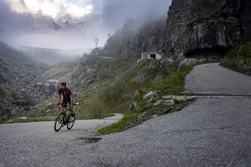 Ciclista su strada di montagna vicino a Camere d'Aria a Locana, Italia, con picchi drammatici e valle nebbiosa sullo sfondo