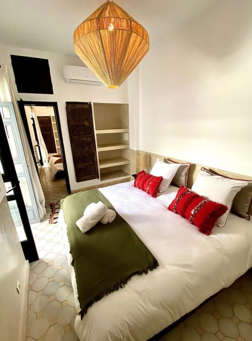 Chambre au Riad 191 à Marrakech avec lit blanc, coussins rouges, luminaire suspendu tissé et étagères intégrées